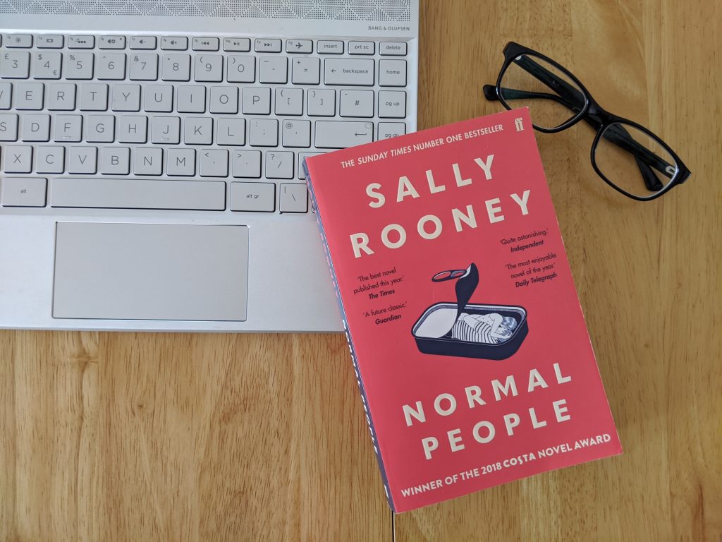 The book Normal People rests on a desk next to a silver laptop and a pair of black glasses