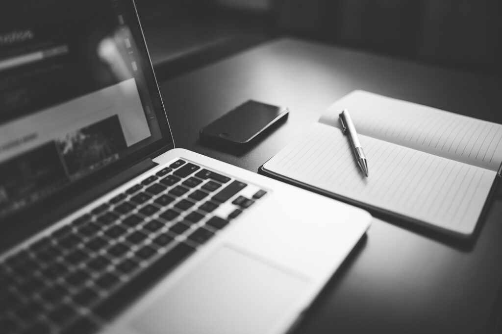 A laptop, mobile phone, notebook and pen on a table to illustrate my writing and editing portfolio page. The image is black and white